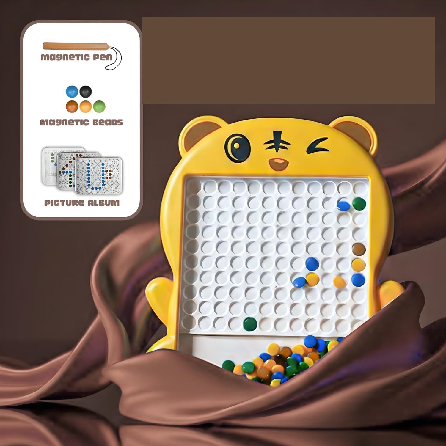 A yellow bear-shaped magnetic bead board toy with a white grid and colorful magnetic beads, shown with an inset card listing included items: a magnetic pen, magnetic beads, and a picture album. The toy is set against a dark brown background with draped brown fabric.